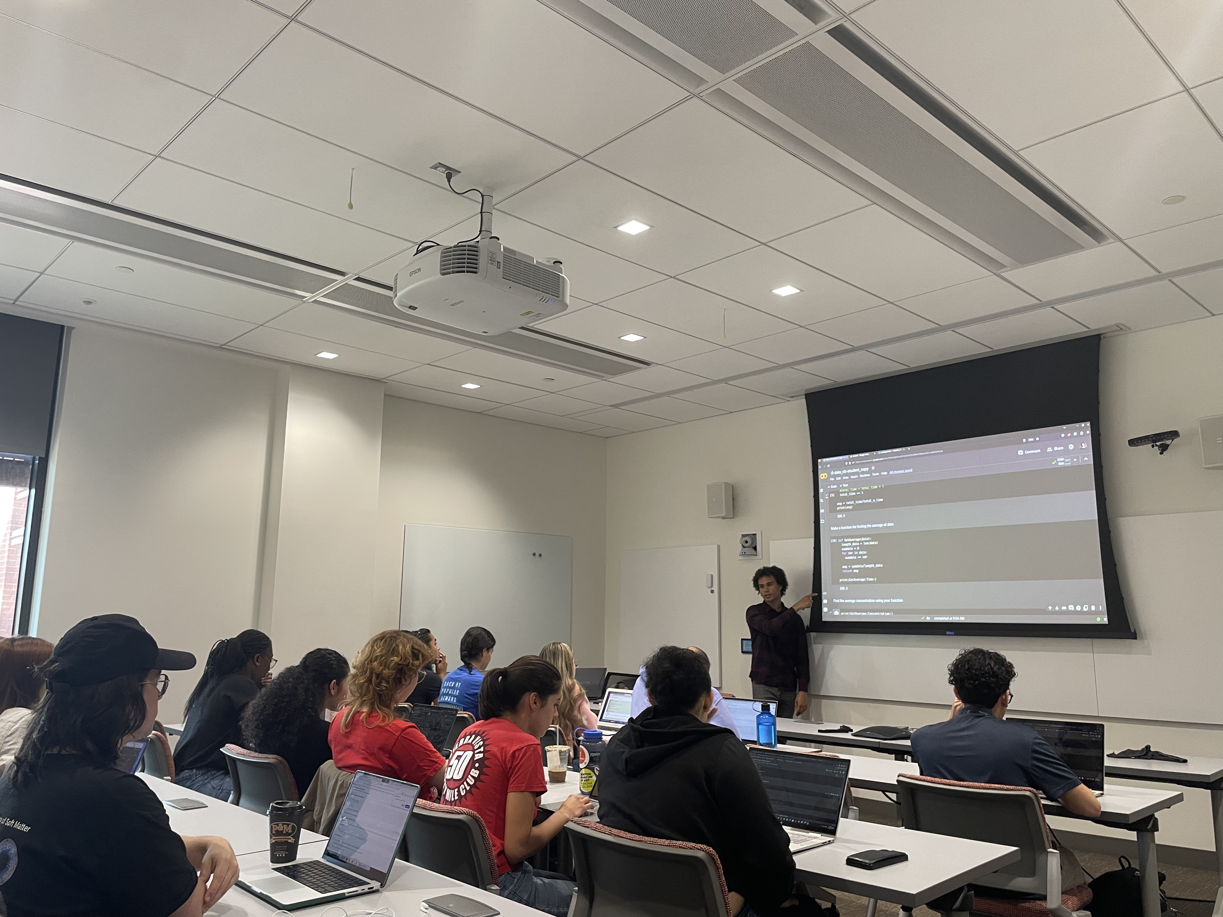 REU student taking notes during a Python lecture with a PEB graduate student at the front of the room teaching.