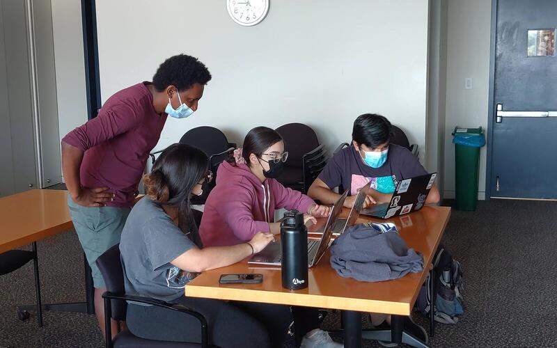 Group of 4 students talk while looking at their computers