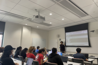 REU student taking notes during a Python lecture with a PEB graduate student at the front of the room teaching.