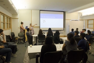 Two presenters point to a slide during a powerpoint presentation in front of a large group