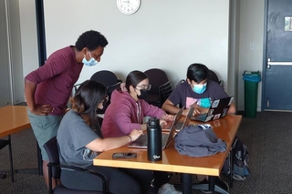 Group of 4 students talk while looking at their computers