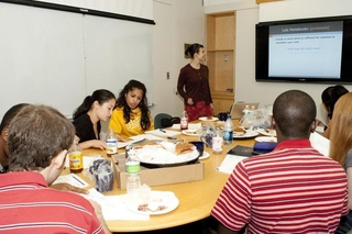 Students sitting around a large table while one presents a powerpoint at the head of the table