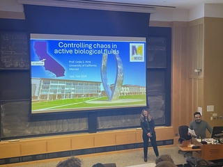 Prof. Linda Hirst standing in front of her presentation while Charles Lomba introduces her